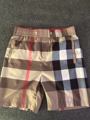 Beige rutiga badbyxor med resårmidja - Snygga badbyxor i beige med klassiskt rutigt mönster i svart, vitt, blått och rött. Shortsen har resår i midjan och sidofickor samt en bakficka. Perfekta för sommaren och har en avslappnad passform.saknar band för att dra åt.