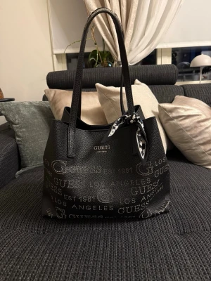 Svart handväska från Guess - Snygg svart handväska från Guess med silverfärgad logotyp och texttryck över hela väskan. Väskan har två långa handtag och en dekorativ svartvit scarf knuten på ena sidan. Tillverkad i strukturerat konstläder med rymlig insida.