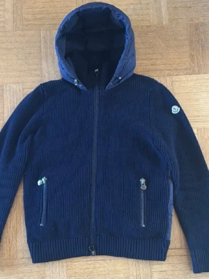 Moncler Cardigan  - Säljer en marinblå sällsynt Moncler  i storlek M med stickade ärmar och dunvadderad rygg och huva. Den är i fint begagnat skick och perfekt för vår och höst. Eftersom jag köpt den second hand och saknar kvitto kan jag inte garantera äktheten, och säljer den därför billigt i befintligt skick. Skriv för fler bilder, jag fraktar snabbt!