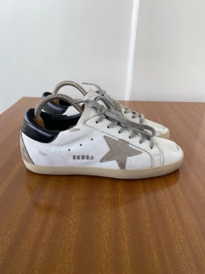 Golden Goose vita sneakers med stjärna - Golden Goose sneakers i vitt läder med grå mockastjärna på sidan, grå skosnören och svart hälparti. Klassisk låg modell med slitna detaljer och beige sula. Perfekta för en avslappnad streetstyle-look. OBS utan kartong