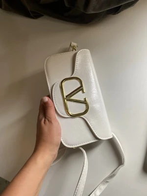 Vit axelremsväska i konstläder - Snygg vit handväska med axelrem i konstläder. Väskan har en enkel, stilren design med guldfärgade detaljer och magnetlås. Perfekt storlek för det viktigaste och lätt att matcha till olika outfits.