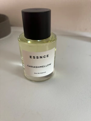 ESSNCE Carashmellow Eau de Parfum - En stilren parfymflaska från ESSNCE med doften Carashmellow. Flaskan är genomskinlig med en svart cylinderformad kork och vit etikett med svart text. Innehållet är ljust gult och flaskan har en modern, minimalistisk design.