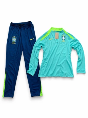 Nike Brasil träningsoverall blå/turkos - Säljer en Brasilien träningsoverall från Nike med långärmad turkos tröja och blå byxor. Tröjan har limegröna detaljer och halv dragkedja, byxorna har elastisk midja och limegröna inslag. Perfekt för fotbollsträning eller sportaktiviteter.