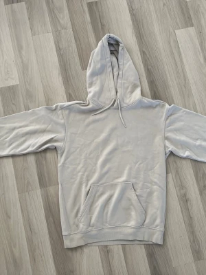 Ljusbeige basic hoodie - En stilren ljusgrå hoodie med klassisk huva och känguruficka framtill. Hoodien har ribbade muddar vid ärmslut och nederkant samt dragsko i huvan. Perfekt för en avslappnad och enkel look. Materialet känns mjukt och bekvämt mot huden.