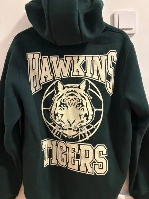 Stranger Things Mörkgrön Hawkins Tigers hoodie  - Snygg mörkgrön hoodie med stor Hawkins Tigers-tryck på ryggen och mindre tryck framtill. Klassisk känguruficka och huva. Perfekt för dig som gillar retrostil och Stranger Things-vibbar. Mjuk bomullsblandning och avslappnad passform.