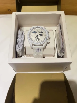 Vit Omega x Swatch Speedmaster klocka - Säljer en vit Omega x Swatch Speedmaster klocka med vit textilrem och blå detaljer. Klockan har tachymeterskala på bezeln och flera subdials på urtavlan. Speedmaster-logga broderad på remmen och unik design inspirerad av rymden. Kommer i originalförpackning. Använd några gånger och haft den i endast 1 dag
