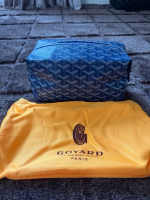Blå necessär  - Snygg blå necessär  med klassiskt mönster i vitt och silver. Rektangulär form med dragkedja upptill och detaljer i blått läder. Kommer med original gul dustbag . Perfekt för att organisera dina småsaker.