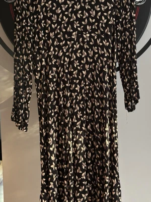 Svart blommig klänning KappAhl - Svart långärmad klänning från KappAhl med vitt blommönster. Klänningen har rund halsringning och dragkedja i ryggen. Materialet är lätt och faller snyggt, perfekt för dig som gillar en stilren men ändå lekfull look.