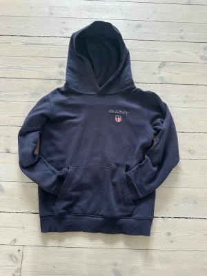 Mörkblå hoodie från GANT - Mörkblå hoodie från GANT med broderad logga och sköld på bröstet. Klassisk känguruficka framtill och ribbade muddar vid ärmslut och nederkant. Tillverkad i mjukt bomullsmaterial och har en stor huva för extra komfort.