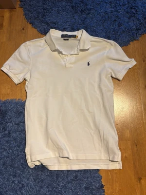 Vit pikétröja från Polo Ralph Lauren - Klassisk vit pikétröja från Ralph Lauren med korta ärmar och krage. Tröjan har två knappar vid halsen och den ikoniska broderade loggan i marinblått på bröstet. Tillverkad i mjuk bomull som känns skön mot huden. Är L i barn men passar XS vuxen storlek.