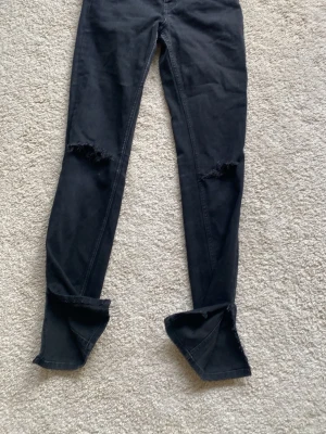 Svarta skinny jeans med hål - Säljer ett par svarta skinny jeans med slitna hål vid knäna och råa kanter vid bensluten. Jeansen har en tight passform och är perfekta för en trendig och avslappnad stil. Materialet är stretchigt denim för extra komfort.