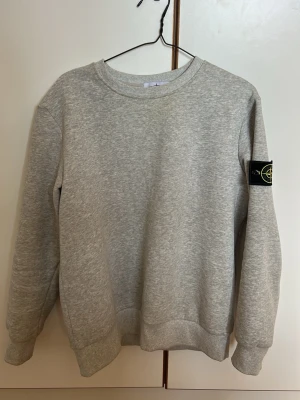 Grå sweatshirt från Stone Island - Säljer en stilren grå sweatshirt från Stone Island med klassisk rund halsringning och ribbade muddar. Tröjan har den ikoniska Stone Island-loggan på vänster ärm. Perfekt för dig som gillar streetwear och enkel design.