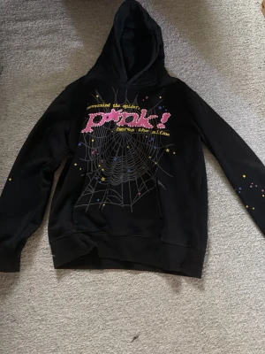 Svart hoodie från Spüer med tryck - Svart hoodie från Spüer med färgglatt tryck på bröstet där det står 'pink!' och ett spindelnätsmotiv. Texten 'nevermind the spider, here's the slime' och små stjärnor i olika färger. Tillverkad i 100% bomull och har klassisk huva.
