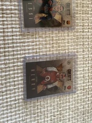 Samlarbilder fotboll Topps Greets - Två samlarkort från Topps Greets-serien med fotbollsspelarna Lionel Messi och Thierry Henry. Korten har färgstarka porträtt och klubbmärken för Barcelona och Arsenal. De är inplastade i genomskinliga skyddsfodral och har detaljerad information på baksidan.
