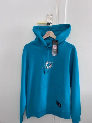 Turkos Miami Dolphins hoodie NFL - Snygg turkos hoodie från BOSS med Miami Dolphins NFL-logga på bröstet och stor text på ryggen. Klassisk känguruficka framtill, justerbar huva med snören och mjukt material. Perfekt för dig som gillar sportig stil och vill sticka ut.