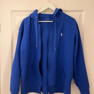 Blå hoodie från Polo Ralph Lauren - Säljer en blå hoodie från Polo Ralph Lauren med dragkedja och huva. Tröjan har två fickor framtill och klassisk logga broderad på bröstet. Perfekt för en avslappnad stil. Använd bara 2-3 gånger, helt ny.