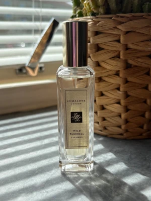 Jo Malone Wild Bluebell Cologne - En elegant parfym från Jo Malone, Wild Bluebell Cologne, i en genomskinlig glasflaska med silverfärgat lock. Doften är fräsch och blommig, perfekt för dig som gillar lätta och naturliga toner. Flaskan har en stilren, rektangulär form och exklusiv etikett. Endast testad 