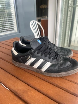 Adidas Samba svart/vit sneaker - Klassiska Adidas Samba sneakers i svart läder med vita detaljer och de ikoniska tre ränderna på sidan. Skon har brun gummisula och svart snörning. Märket syns tydligt på plösen och sidan. Perfekt för dig som gillar retrostil och streetwear.