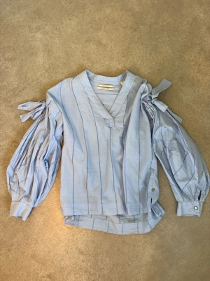 Ljusblå blus med knytärmar  -  ljusblå blus från Second Female med tunna vertikala ränder. Blusen har långa puffärmar med knytband vid axlarna och manschetter med knapp. Jättefin söt blues topp. Använd max två gånger jättefint i sommar. Skriv om du har några funderingar❤️