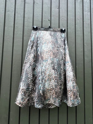 Mönstrad långkjol med ormskinnslook - Säljer en flowy långkjol med coolt ormskinnsmönster i svart, beige och turkos. Kjolen har en lätt utsvängd form och är tillverkad i ett mjukt, glansigt material som ger en snygg siluett. Perfekt för dig som vill sticka ut med ett djurmönster.