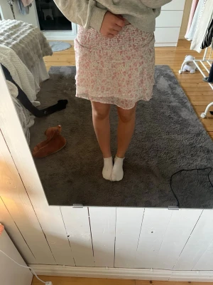 Blommig kortkjol från Gina Tricot - Supersöt kortkjol från Gina Tricot med blommigt mönster i rosa och grönt på vit botten. Kjolen har smock i midjan och volangkant nedtill för en extra feminin touch. Perfekt för vår och sommar!