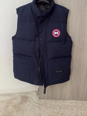 Canada Goose väst - Snygg mörkblå dunväst från Canada Goose med hög krage, två stora framfickor och klassisk logotyp på bröstet. Västen har tryckknappar och dragkedja framtill samt diskret Canada Goose-märke på ena fickan. Perfekt för lager-på-lager under kyliga dagar. Västen är mycket bra skick, använd bara en gång. 