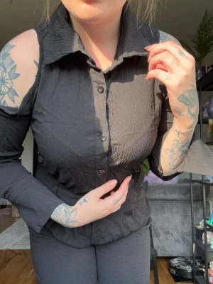 Svart skjorta med cut out och krage - Säljer en svart skjorta med cut out-detaljer på axlarna och klassisk krage. Skjortan har knappar framtill och är tillverkad i ett lätt, skrynkligt tyg. Långärmad modell med unik design som sticker ut. Går till naveln. Endast testad, tagen bild ej använd 