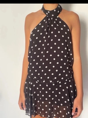 Prickig halterneck klänning - Snygg klänning med vita prickar och öppen rygg. Halterneck-modell som knyts i nacken och har ett flowy, luftigt tyg. Perfekt för dig som vill ha en trendig och bekväm look med lite extra edge. Kort modell som passar till varma dagar.