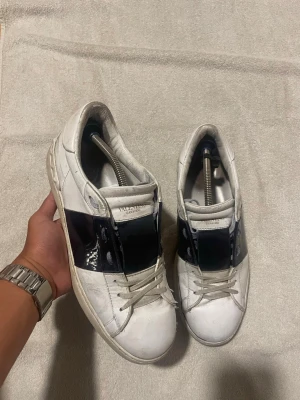 Valentino opens - Säljer ett par vita sneakers från Valentino med en bred svart glansig remsa över mitten. Skorna har klassisk låg profil, vita snören och diskret logga på plösen. Tillverkade i läder med gummisula. Perfekta för dig som gillar stilrena men ändå unika detaljer.