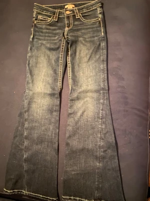 Mörkblå bootcut jeans med kontrastsömmar - Säljer ett par mörkblå bootcut jeans med snygga kontrastsömmar och klassisk femficksmodell. Jeansen har lätt slitna detaljer och utsvängda ben, perfekta för en retroinspirerad look. Materialet är denim i bomull med lite stretch för skön passform. Oanvända och köptes för 500.