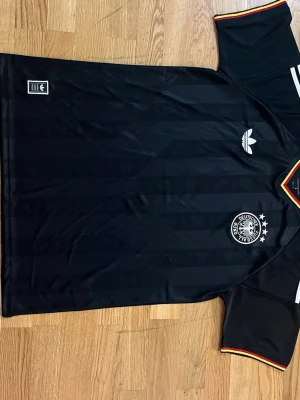 Svart Tyskland fotbollströja Adidas - Snygg svart fotbollströja från Adidas med Tysklands landslagsemblem och fyra stjärnor på bröstet. Tröjan har diskreta ränder, Adidas-logga och detaljer i rött, gult och svart vid kragen och ärmsluten. Perfekt för fotboll eller streetwear.    Fraktar samma eler dagen efter du köpt!!! Endast en kvar i lager!!!