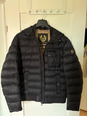 Svart pufferjacka från Belstaff - Snygg svart pufferjacka från Belstaff med huva och dragkedja framtill. Jackan har en bröstficka med knappar, två sidofickor och ribbade muddar. Märkeslogga på ärmen och insidan. Perfekt för kalla vinterdagar. Storlek: UK 40 / IT 50 / US 40 (motsvarar ca M–L herr). Mycket bra skick!