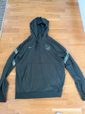 Mörkgrön Nike Atlético hoodie - Snygg mörkgrön hoodie från Nike med Atlético de Madrid-logga på bröstet och halv dragkedja. Hoodien har dragsko i huvan, ribbade muddar och en stor magficka. Perfekt för dig som gillar sportig stil och vill representera ditt favoritlag.