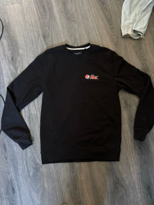 Svart sweatshirt från Berkeley - Svart sweatshirt från Berkeley med liten Pepsi Max-logga på bröstet. Klassisk rund halsringning och långa ärmar. Tillverkad i mjuk bomullsmix som är skön att ha på sig. Perfekt för en chill och stilren look.