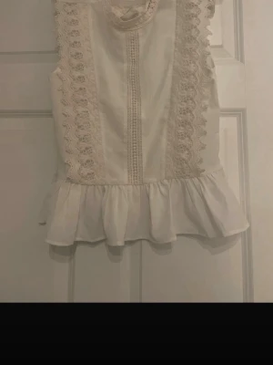 Vit blus  - Söt vit blus med volang nedtill och fina spetsdetaljer framtill. Blusen är ärmlös och har en rund halsringning samt dekorativa sömmar. Perfekt för en fräsch och romantisk look. Materialet känns lätt och luftigt. Aldrig använd 
