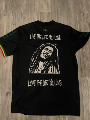 Svart Bob Marley x Ajax t-shirt - Unik svart t-shirt med Bob Marley-tryck och texten 'Live the life you love, love the life you live' på framsidan. Baksidan har Ajax-logga, Ziggo, Adidas och färgglada detaljer i rött, gult och grönt. Kortärmad och tillverkad i bomull.