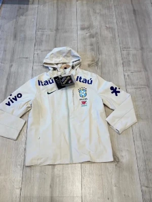 Vit Brasil Nike regnjacka Gore-Tex - Snygg vit regnjacka från Nike med Brasilien-logga och flera sponsortryck i blått. Jackan har huva med dragsko, hel dragkedja och är tillverkad i slitstarkt Gore-Tex-material. Perfekt för dig som gillar sportig stil och vill hålla dig torr.Den är L men passar mer som M