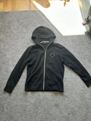 Svart zip hoodie från BOSS - Snygg svart hoodie från BOSS med dragkedja framtill och huva. Diskret BOSS-logga på bröstet i vitt. Tillverkad i mjukt bomullsmaterial som känns skönt mot huden. Perfekt för en avslappnad och stilren look.