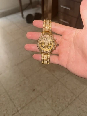 Guldig klocka med stenar - Säljer en guldig klocka med glittriga stenar på både armband och urtavla. Vissa stenar har ramlat av dock. Klockan har en rund form och detaljerad design med flera små stenar som ger en lyxig känsla. Perfekt accessoar för dig som vill sticka ut och glänsa.