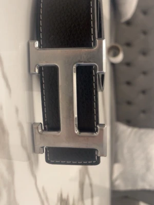 Svart bälte med stor silverspänne - Snyggt svart bälte i skinn med ett stort, rektangulärt silverspänne format som ett H. Bältet har vita sömmar längs kanterna som ger en schysst kontrast. Perfekt accessoar för att lyfta din outfit. 110cm