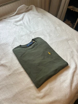 Olivgrön t-shirt från Polo Ralph Lauren - Snygg olivgrön t-shirt från Polo Ralph Lauren med klassisk gul broderad logga på bröstet. T-shirten har rund hals och är kortärmad, perfekt till en avslappnad stil. Tillverkad i mjuk bomull för maximal komfort.