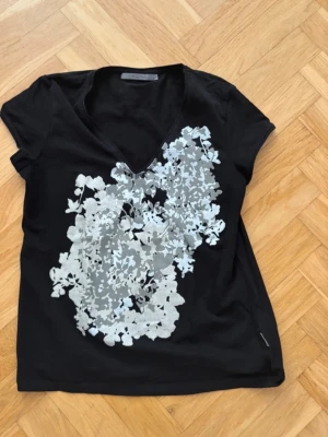 Svart t-shirt med blommigt tryck - Snygg svart t-shirt med v-ringning och stort blommigt tryck i silver och grått framtill. Kortärmad modell i mjukt bomullsmaterial som känns skön mot huden. Perfekt för dig som vill ha en t-shirt med lite extra detaljer.