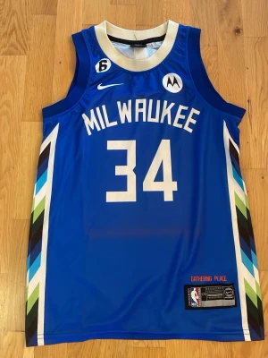 Blå Milwaukee Bucks basketlinne - Snyggt blått basketlinne med nummer 34 och texten MILWAUKEE på framsidan samt ANTETOKOUNMPO på ryggen. Linnet har vita och färgglada detaljer längs sidorna och är tillverkat i ett lätt, ventilerande material. Perfekt för basketträning eller streetwear.storlek 176. Okej skick är lite färgblekt på ryggen men det syns knappast.