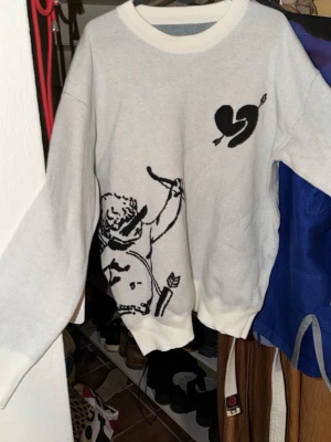 Vit tröja med tryck och text - Vit oversized tröja med svart tryck av en amor med pilbåge på framsidan och ett trasigt hjärta. På baksidan står texten 'I'd rather have loyalty than love' i svart. Tröjan har ribbade muddar och rund hals. Perfekt för dig som gillar statement-plagg.