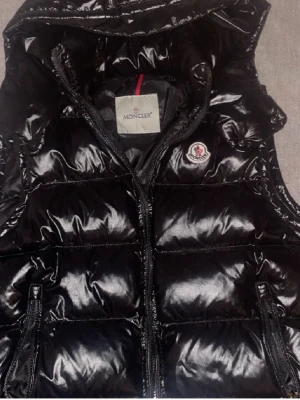 Svart glansig dunväst från Moncler - Snygg svart dunväst från Moncler med glansig finish, hög krage och dragkedja framtill. Västen har två fickor med dragkedja och klassisk Moncler-logga på bröstet. Perfekt för lager-på-lager och streetstyle.