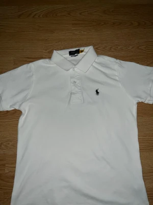 Vit pikétröja från Polo Ralph Lauren - Klassisk vit pikétröja från Polo Ralph Lauren med broderad logga på bröstet. Tröjan har krage, knappar framtill och korta ärmar. Perfekt för en stilren och tidlös look.