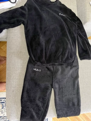 Svart fluffigt loungewear-set - Mysigt svart loungewear-set i mjuk fleece med långärmad tröja och matchande byxor. Båda delarna har broderad vit text på bröstet och benet. Perfekt för chill hemma eller när du vill vara extra bekväm.