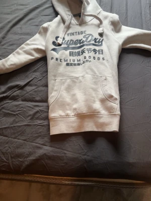 Ljusgrå Superdry hoodie med tryck - Ljusgrå hoodie från Superdry med stort mörkblått tryck på bröstet och japanska tecken. Klassisk känguruficka framtill, ribbade muddar och justerbar huva med snören. Perfekt för en avslappnad streetwear-look.