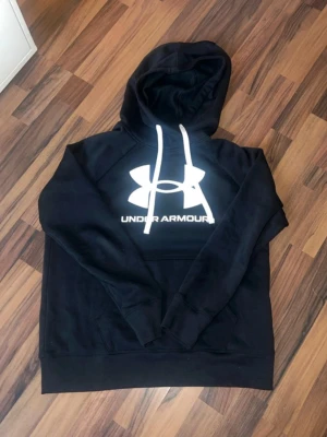 Svart hoodie från Under Armour - Snygg svart hoodie från Under Armour med stor vit logga och text på bröstet. Hoodien har vit dragsko i huvan och en klassisk känguruficka framtill. Tillverkad i mjukt material som passar perfekt för en avslappnad stil.