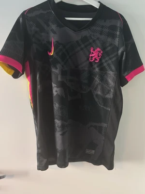 Cole palmer tröja  - ÄKTA! Snygg svart fotbollströja med mönstrad design, rosa och gula detaljer på ärmarna samt rosa lejonemblem på bröstet. V-ringad hals och tryck med namn och nummer 'Palmer 20' på ryggen. Perfekt för dig som älskar fotboll och vill sticka ut på planen.